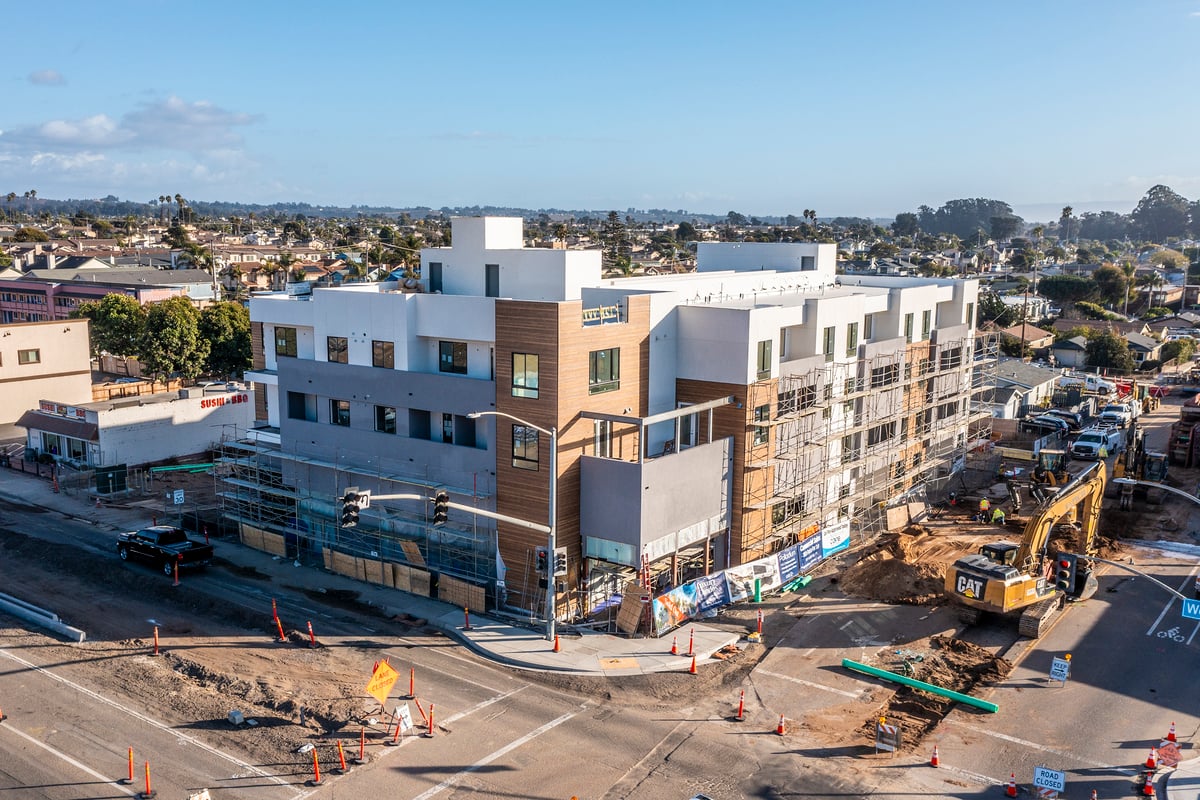 Aerial view of the Palladium development under construction at a bustling intersection in Grover Beach, showing the near-complete modern multistory buildings with workers visible on site and scaffolding along the exterior.